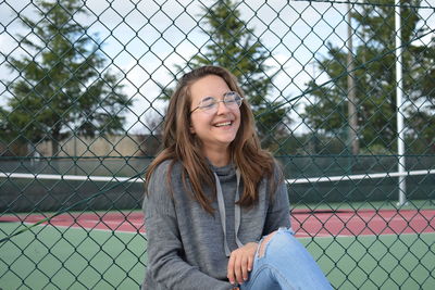 Portrait of smiling young woman standing by chainlink fence