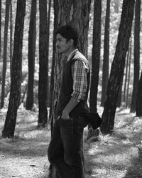 Side view of young man standing by tree trunk in forest