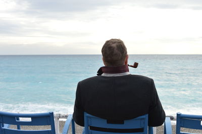 Rear view of man with smoking pipe sitting by sea against sky