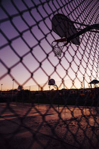 Silhouette basketball hoop seen through chainlink fence against sky during sunset