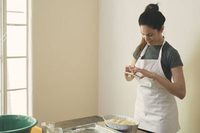 Woman in apron photographing challah bread with smart phone on table against wall