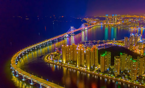 Illuminated bridge over river by buildings against sky at night