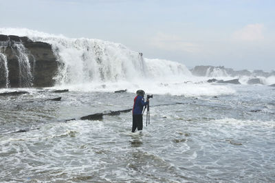 Photographer standing with tripod in sea against waterfall against sky