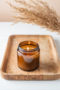 Cozy burning candle in brown glass jar, winter home interior decor on wooden tray with dry reed 