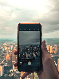 Cropped image of hand holding smart phone against sky in city