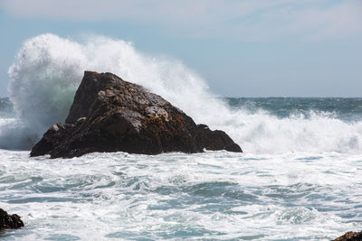 Waves splashing on rocks at shore against sky