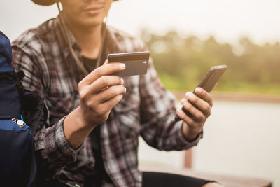 Midsection of young man holding smart phone and credit card while sitting outdoors