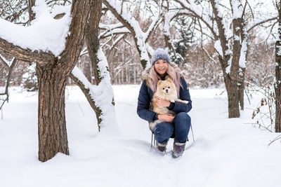 Young woman carrying dog while crouching in snow during winter