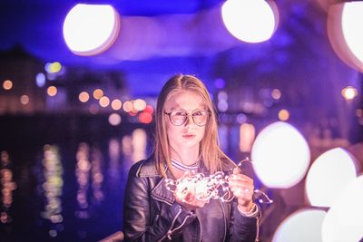 Portrait of young woman wearing eyeglasses holding illuminated string lights in city at night