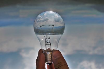 Close-up of hand holding light bulb against sky