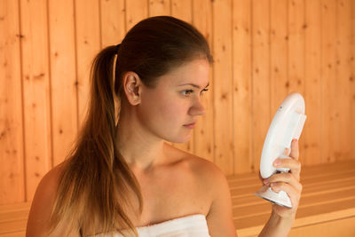 Beautiful young woman holding mirror while sitting in sauna