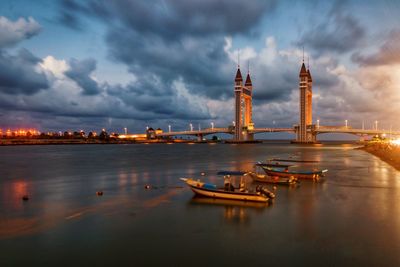 Boats moored in river against dramatic sky and bridge