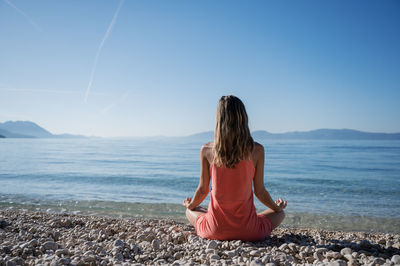 Rear view of woman meditating at seashore against sky