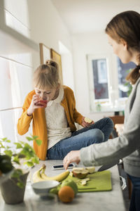 Woman cutting fruits with girl drinking juice on kitchen counter at home