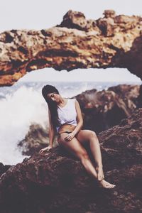 Full length of sensuous woman sitting on rock formation by sea