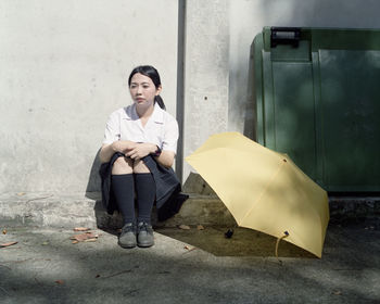 Thoughtful woman sitting by yellow umbrella against wall