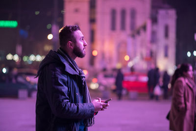 Side view of man holding mobile phone while standing on street in city at night