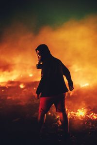 Rear view of young man wearing oxygen mask while standing on field against fire at night