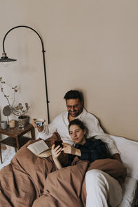 Non-binary person sharing smart phone with boyfriend sitting on bed at home