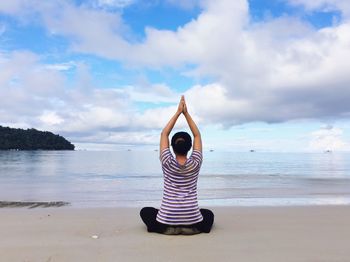 Rear view of woman meditating while sitting on beach against cloudy sky