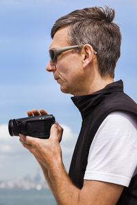 Side view of man holding camera against sky