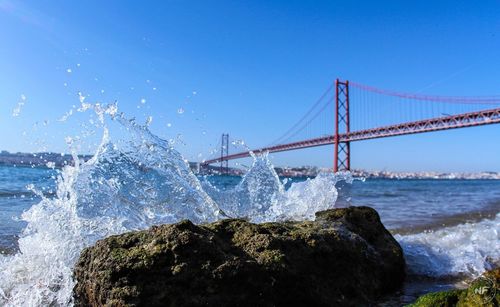 Wave splashing on rock by april 25th bridge against clear blue sky