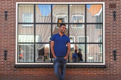 Smiling young man with hands in pockets standing against window of building