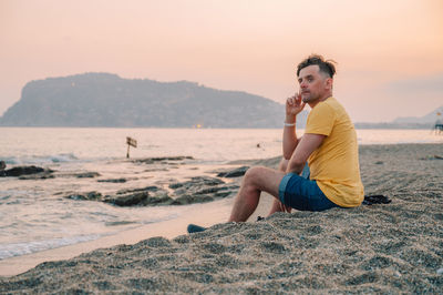 Side view of woman sitting on sand at beach against sky