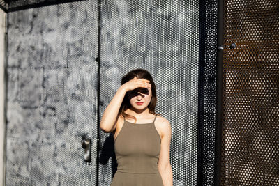 Portrait of young woman shielding eyes against warehouse during sunny day