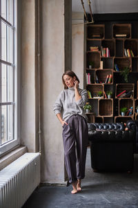 Full length of smiling woman standing by window at home