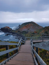 Wooden footbridge leading towards sea against sky