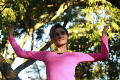 Low angle view of woman looking away with arms raised while standing against trees