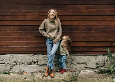 A young laughing mother with a little daughter toddler stand near a wooden wall.