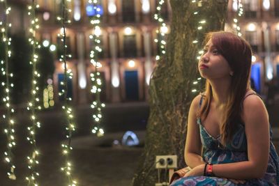 Young woman looking away by illuminated lighting equipment on tree at night