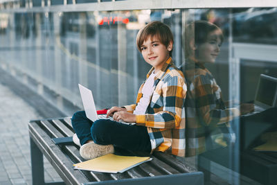 Portrait of boy using laptop while sitting on bench against modern building