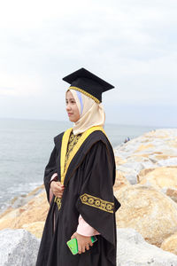 Young woman in graduation gown standing at beach against cloudy sky