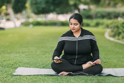 A happy young girl of indian nationality is sitting in a lotus position in the park with a phone 