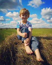 Full length of boy sitting on hay at field against sky