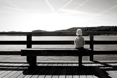 Girl sitting on bench at shore against sky