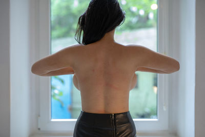 Rear view of shirtless seductive woman standing in front of window at home