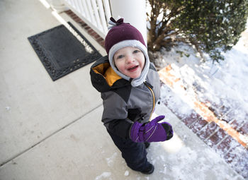 Boy in winter gear standing on snowy front porch looks up at camera