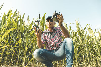 Low angle view of mature man wearing sunglasses holding drone while crouching on field against sky