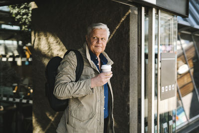 Businessman with backpack holding disposable cup while standing at railroad station entrance