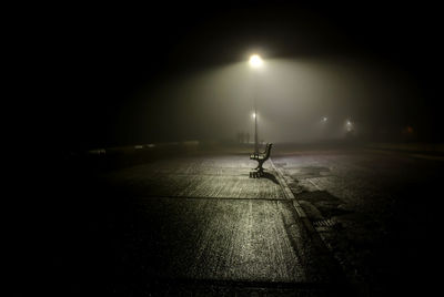 Empty bench on illuminated sidewalk by street at night