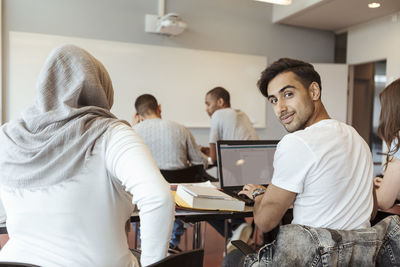 Portrait of smiling man using laptop while sitting with friends in classroom