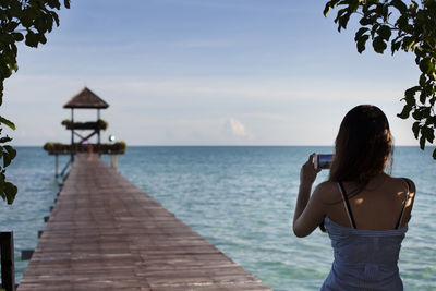 Rear view of woman photographing gazebo over sea against sky