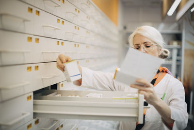 Mature female pharmacist holding prescription paper checking medicine by drawer in store