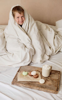 Little boy wrapped in a blanket with breakfast in bed. cocoa with marshmallows and macaroons  tray