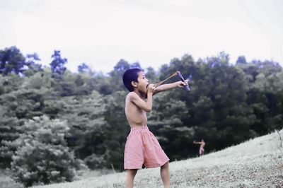 Shirtless boy aiming slingshot while standing on land against trees
