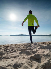 Full length healthy man stretching legs in morning on beach.